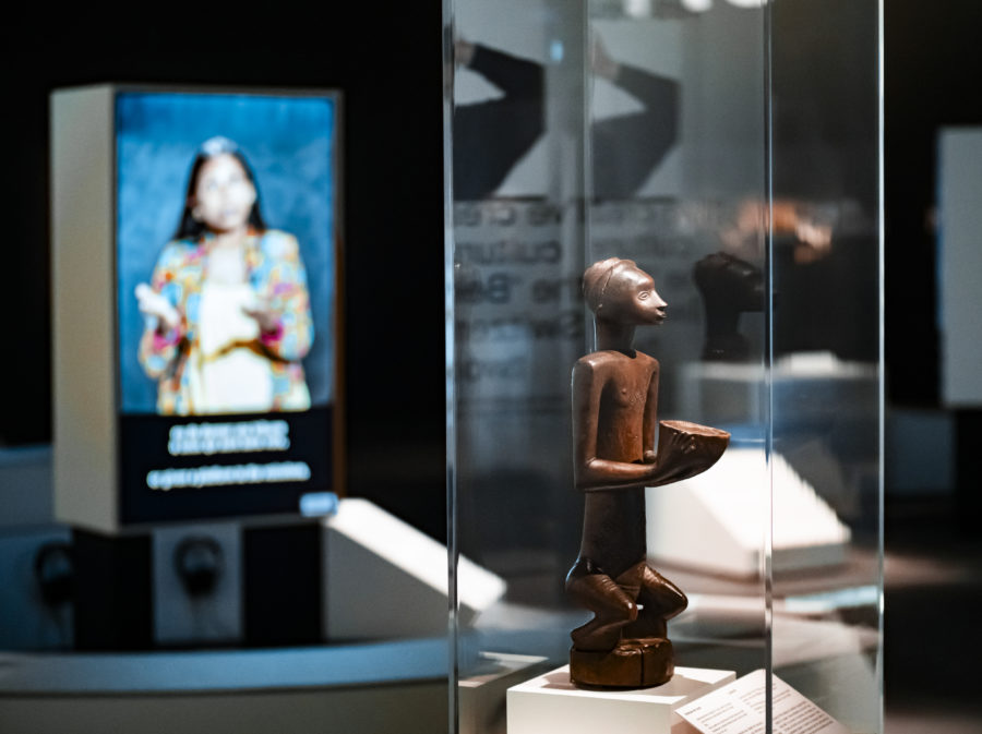 Statue en bois dans une vitrine avec, dans le fond, une personne floue sur un écran MEG