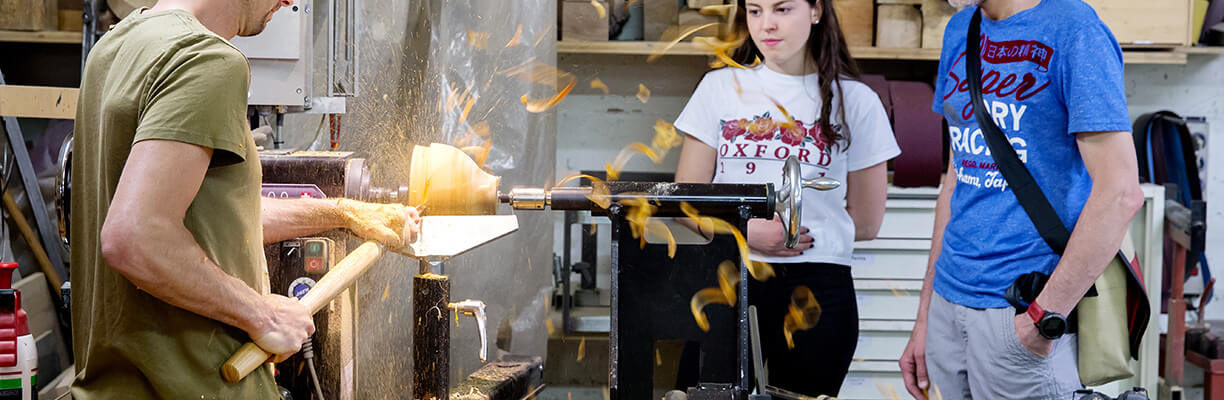 Homme taillant du bois avec un tour à bois. Un homme et une femme le regarde JEMA
