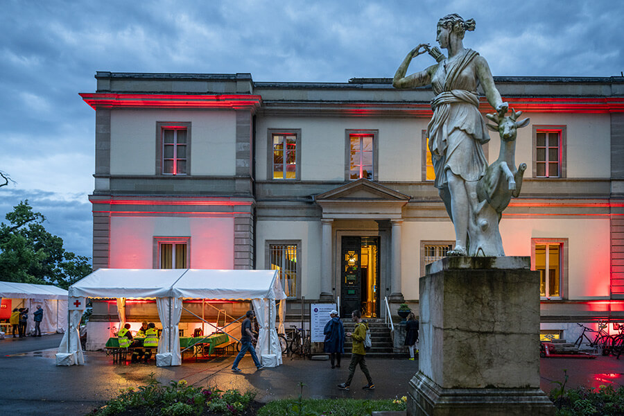 Une statue et des tentes devant la façade du Musée d'histoire des sciences éclairé d'une lumière rouge
