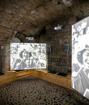 Pièce en sous-sol, trois écrans sur lesquels l'image d'une femme parlant devant des micros est projetée au Musée de la Réforme