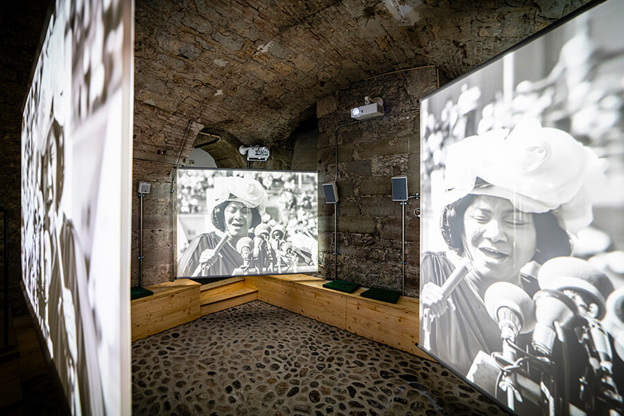 Pièce en sous-sol, trois écrans sur lesquels l'image d'une femme parlant devant des micros est projetée au Musée de la Réforme