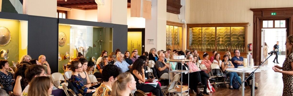 Public assis dans la salle des armures du MAH écoutant une oratrice