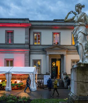 Une statue et des tentes devant la façade du Musée d'histoire des sciences éclairé d'une lumière rouge