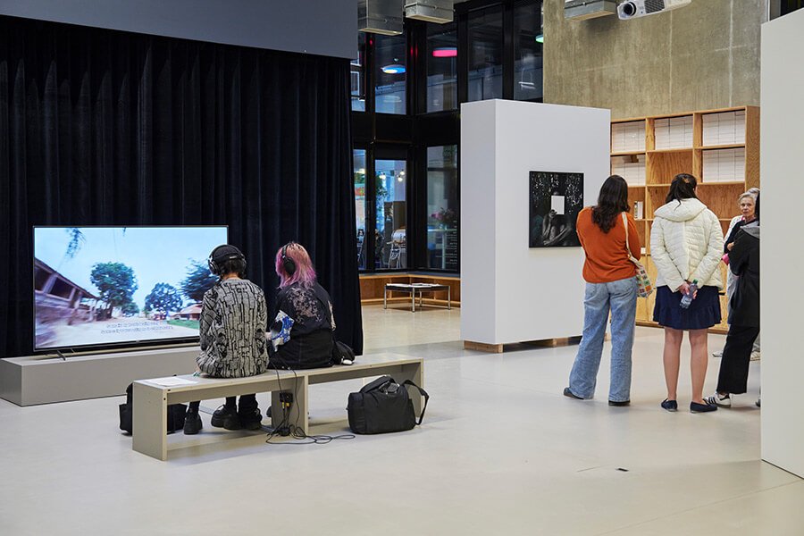 Deux personnes assises sur un banc regardant un écran avec casques sur les oreilles, groupe de personnes debout écoutant une guide au FMAC